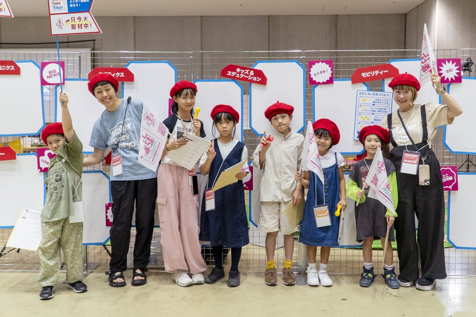 大人と子どもたちが赤いベレー帽をかぶり、旗やマイクを持って並んでいる